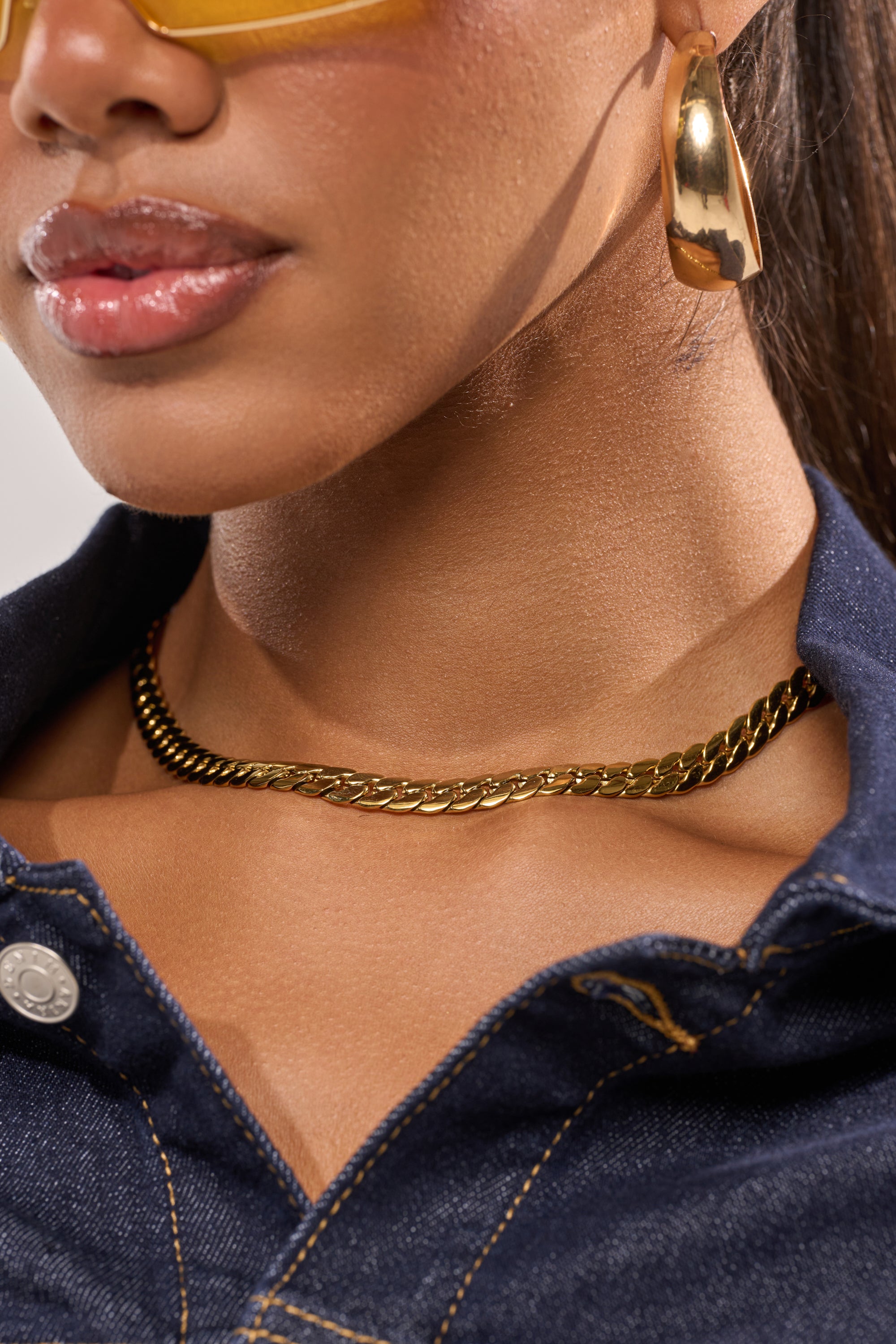 Close-up of a woman in gold hoop earrings, yellow-tinted sunglasses, and a dark denim jacket with visible stitching, styled with the 16 INCH MIAMI CHAIN 8MM gold-tone necklace near the collar. 1
