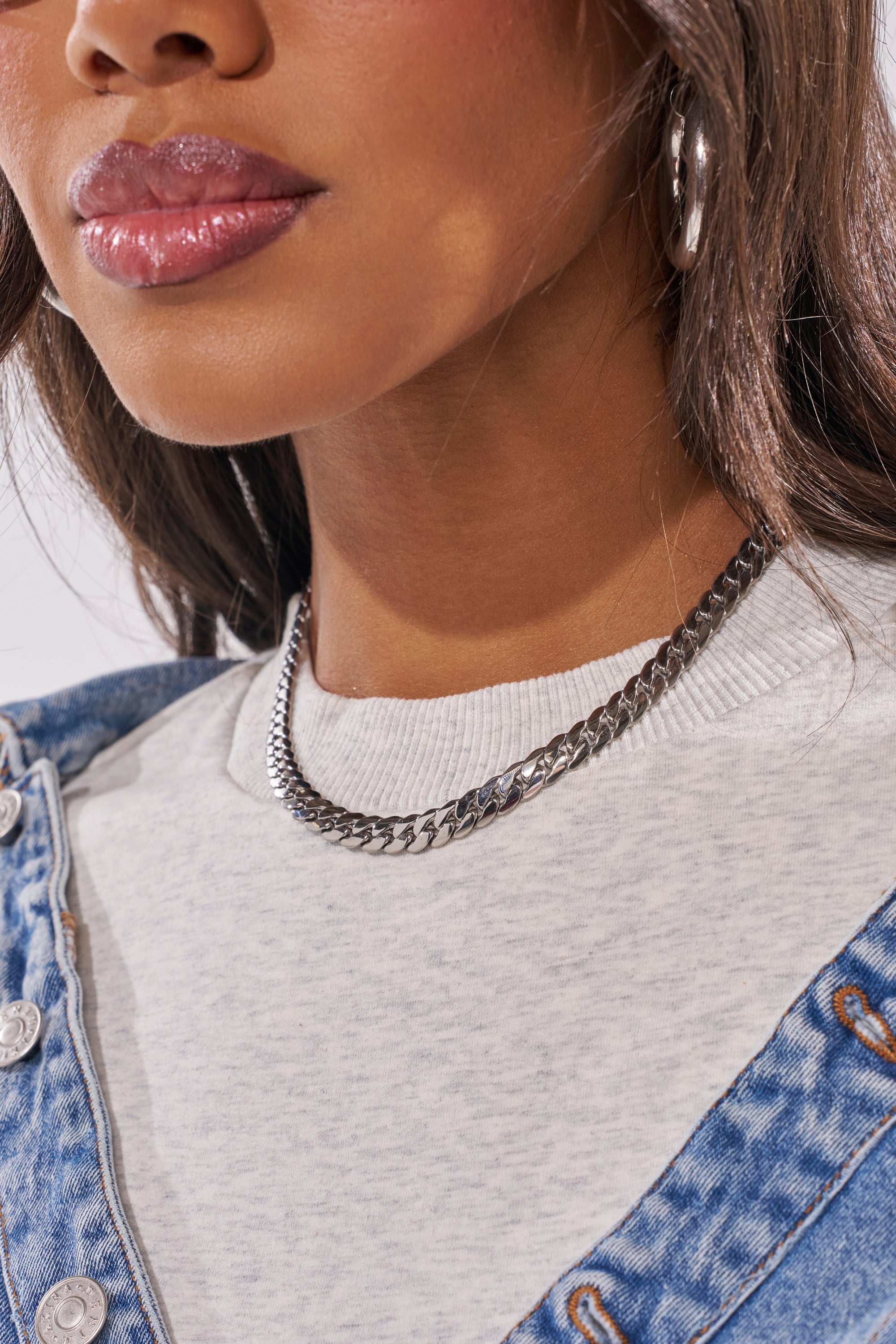 A close-up shows a woman wearing the 16 INCH MIAMI CHAIN 8MM IN SILVER, paired with a white ribbed crew neck shirt, denim jacket, silver hoop earrings, and glossy pink lipstick. Only her lower face and upper chest are visible. 1