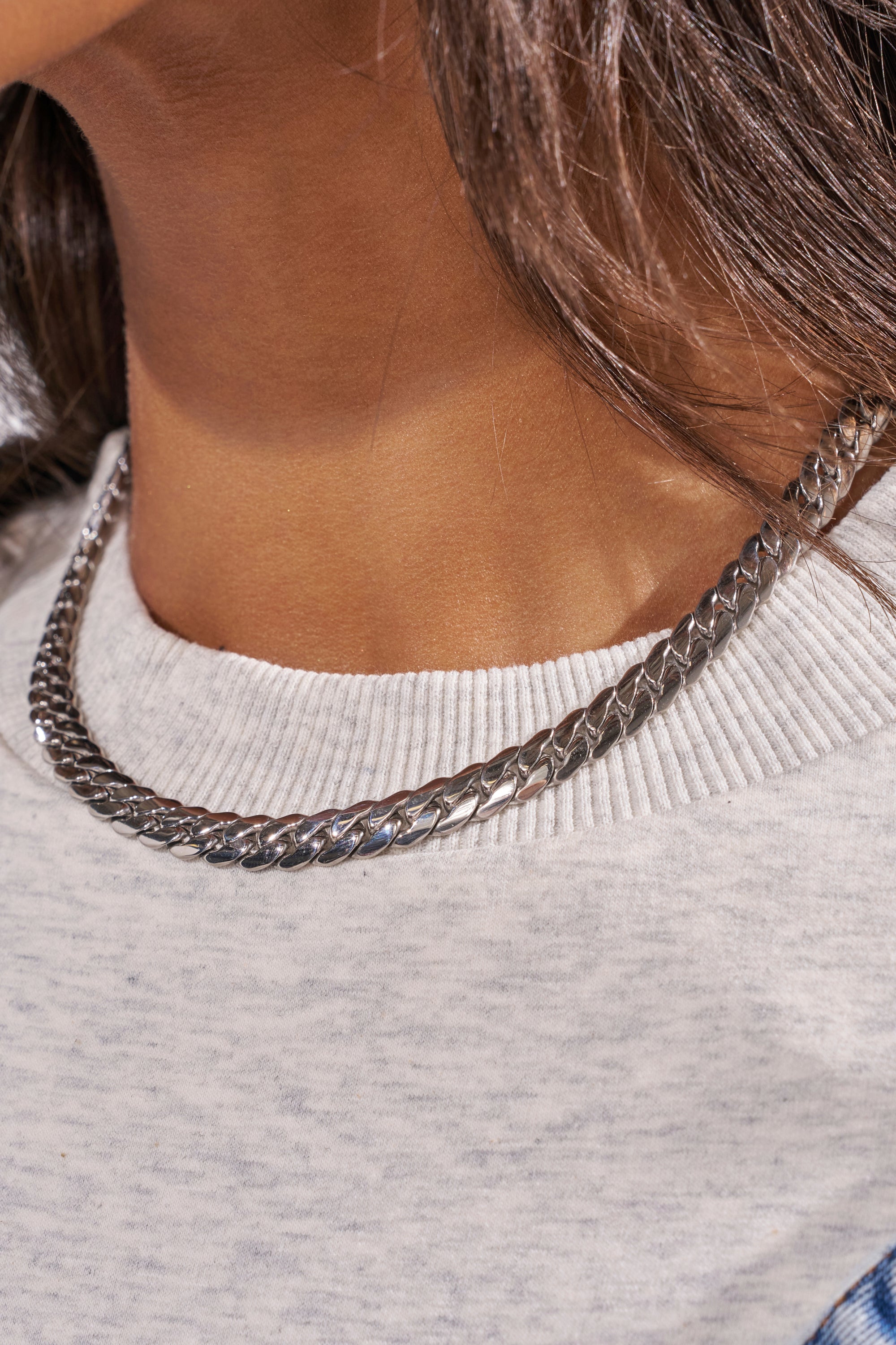 Close-up of a person wearing the 16 INCH MIAMI CHAIN 8MM IN SILVER over a light grey ribbed crew neck shirt. The flat curb-style chain sits along the neckline with brown hair partially visible. 2
