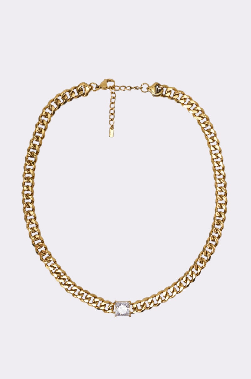 The STAINLESS STEEL SQUARE CZ NECKLACE features a gold-tone Cuban chain with a single square-cut clear gemstone in the center, shown against a plain light background. 0