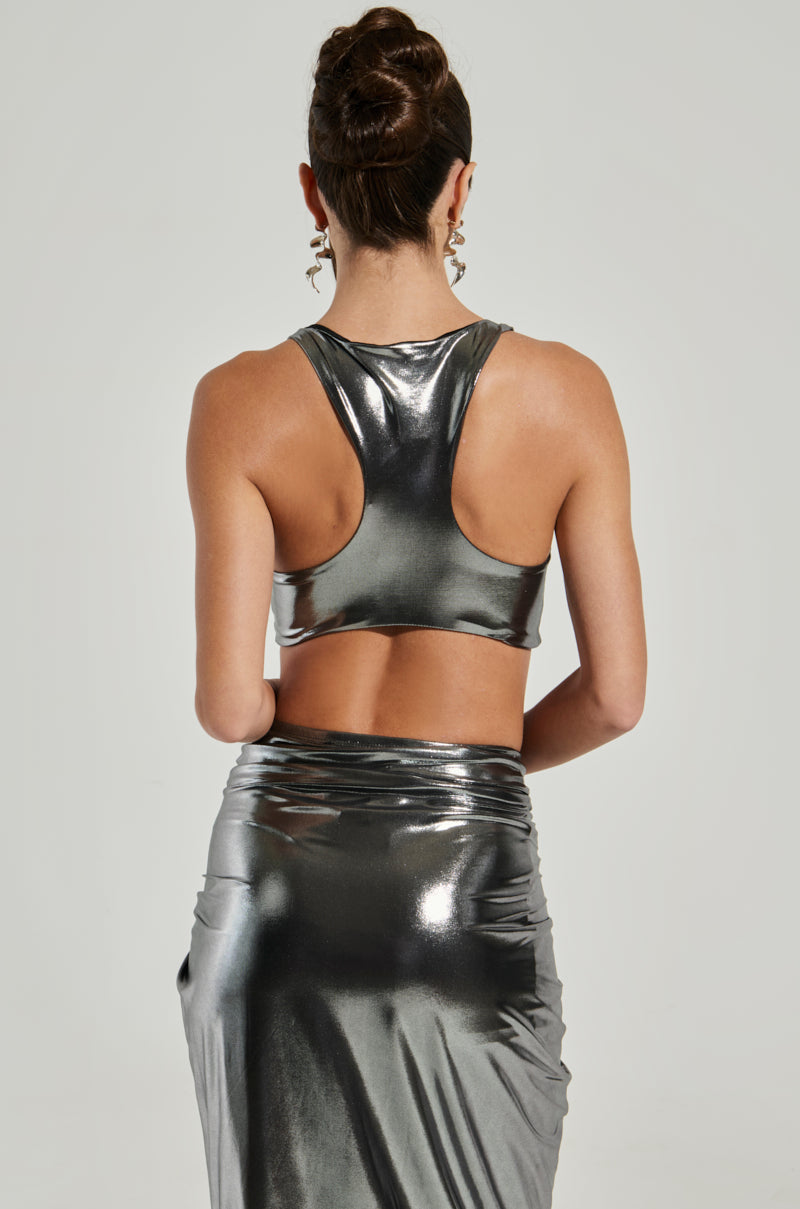 A woman wears the CAN'T STOP MY SHINE TOP with a fitted skirt, standing with her back to the camera to highlight its cropped silhouette. Her sleeveless metallic top perfectly complements her styled bun and large dangling earrings against a plain light-colored background that enhances her outfit's shimmer.