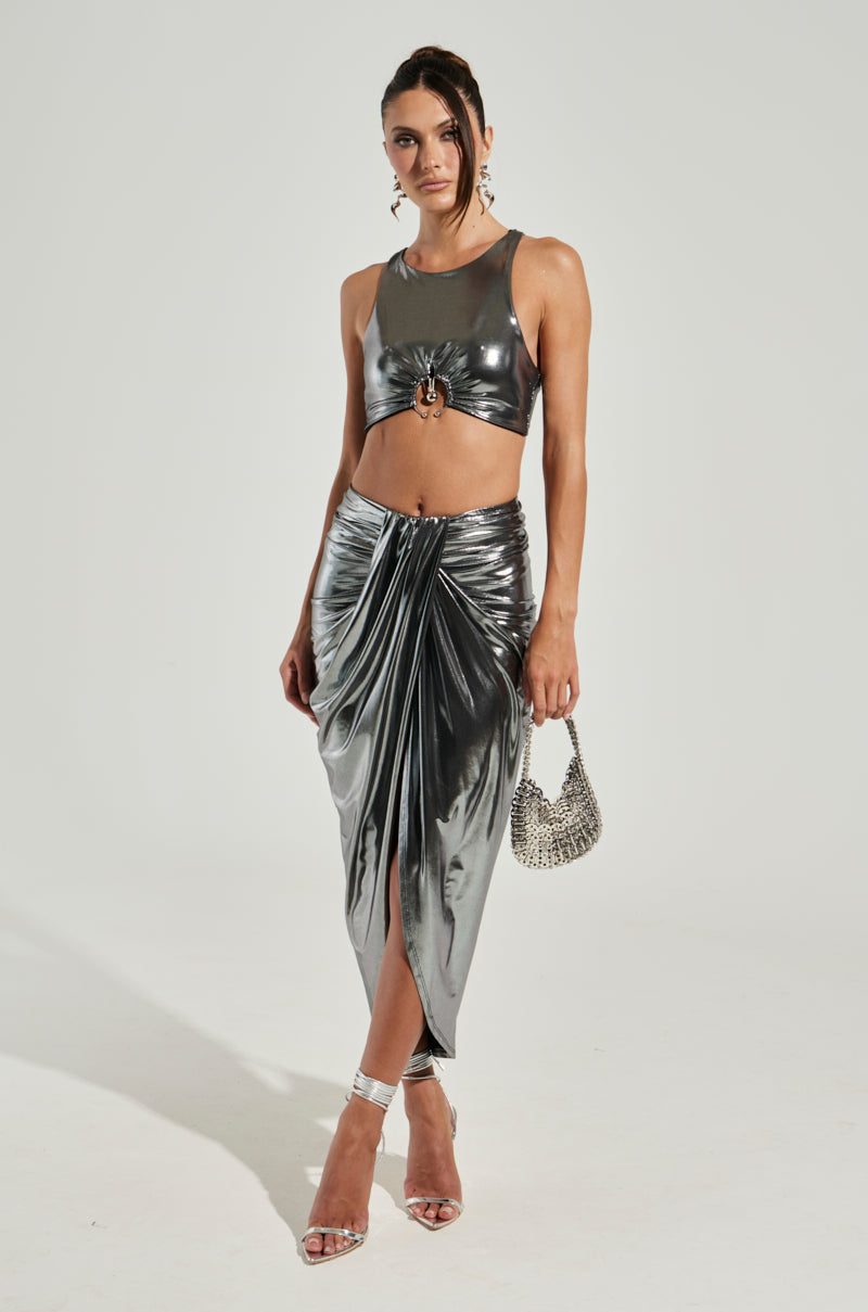 A person stands wearing the CAN'T STOP MY SHINE TOP in a shiny silver cropped silhouette with a draped skirt featuring a knot detail. They hold a small metallic bag in one hand and wear strappy silver high heels. Their hair is styled up, complemented by hoop earrings against a plain white background.