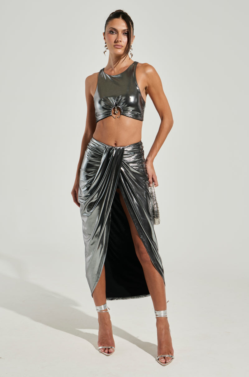 A woman poses in a cropped silhouette wearing the CAN'T STOP MY SHINE TOP, which showcases a shiny silver racerback design. She pairs it with a high-slit skirt, holds a small purse, and wears strappy high heels. Her hair is styled up as she stands against a plain background.