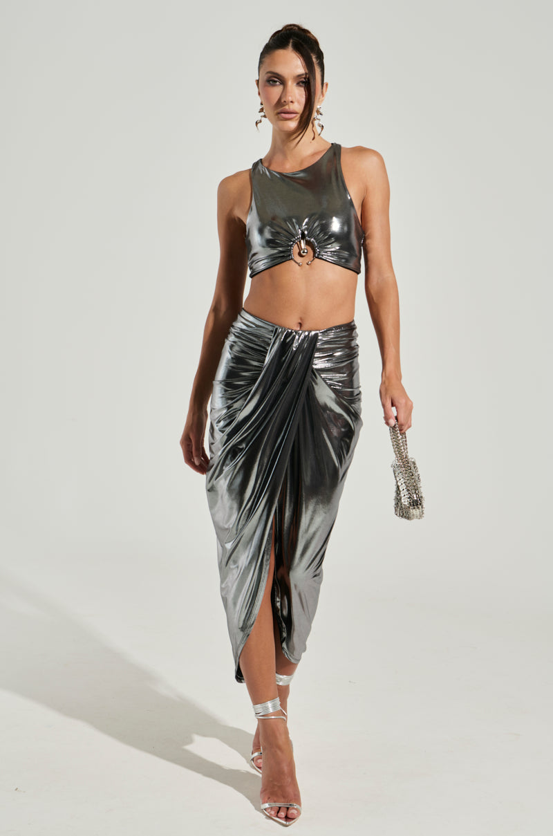 A woman is wearing the CAN'T STOP MY SHINE TOP, a metallic silver cropped silhouette with an ornamental front detail, paired with a matching high-waisted skirt featuring a front slit. She completes her look with a small, shiny purse and silver heels, posing elegantly against a plain background.