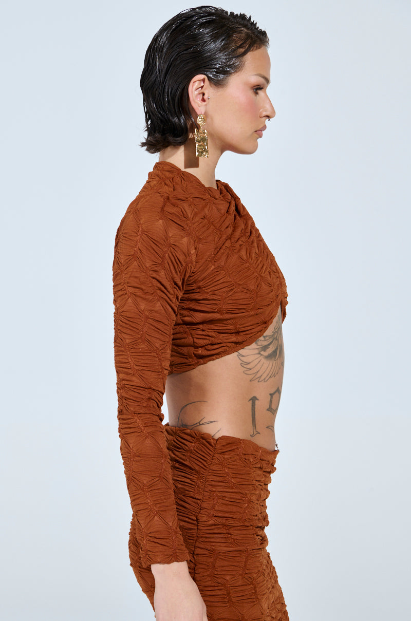 A person with short dark hair and gold earrings is in profile, wearing the MAYBE NEXT TIME Ruched Long Sleeve Top in Brown. An intricate tattoo adorns their side against a plain, light background.