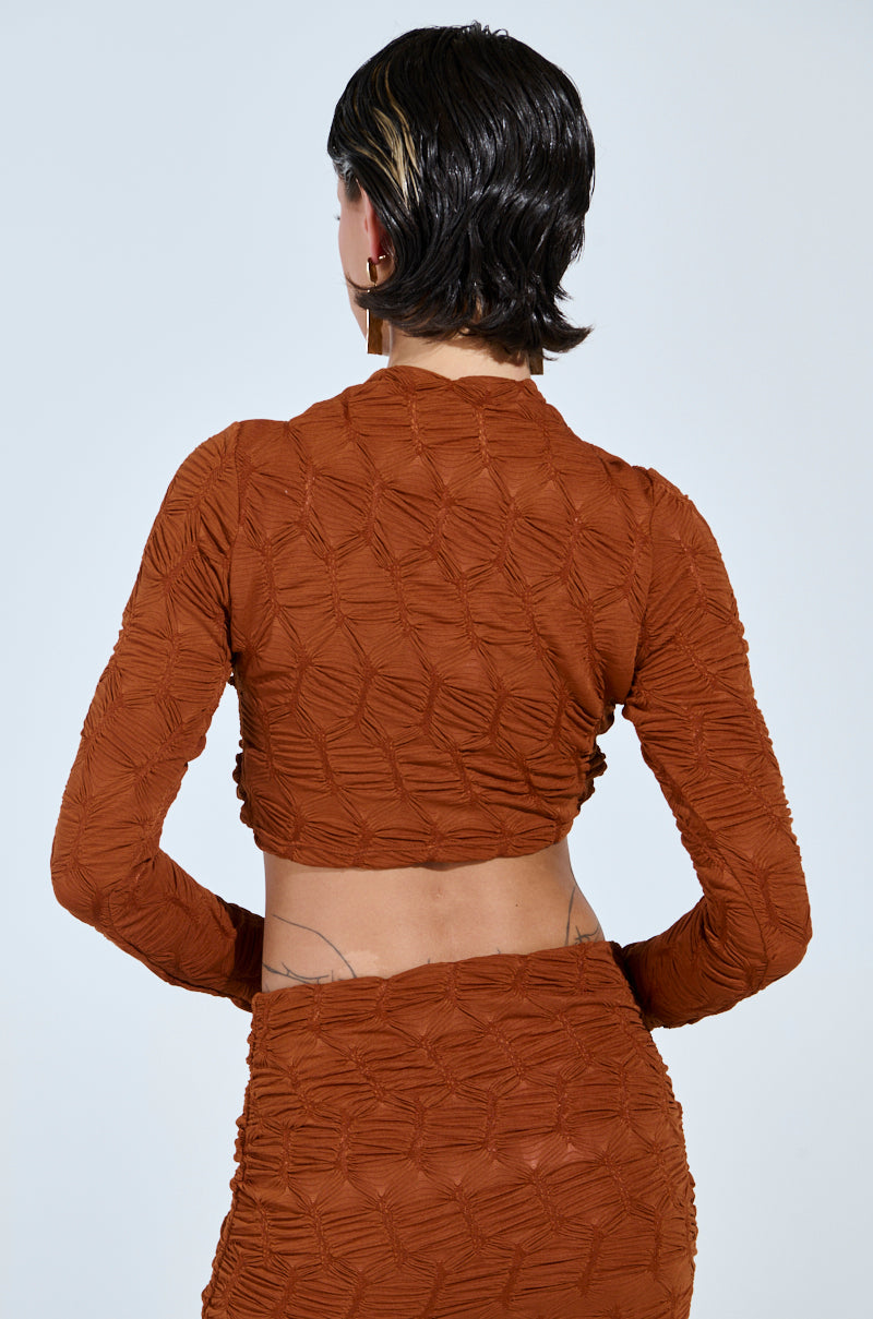 A person with short, dark hair highlighted blonde at the tips turns away from the camera, wearing a textured rust-colored top styled like the "MAYBE NEXT TIME RUCHED LONG SLEEVE TOP IN BROWN" and a matching skirt. Two tattoos adorn their lower back.