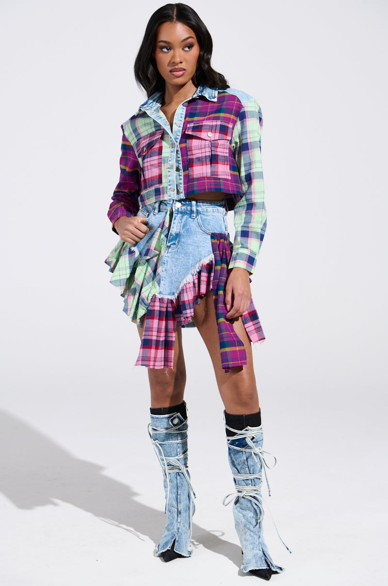 A person wearing a colorful plaid shirt and a THINK ABOUT ME MORE MINI SKIRT WITH PLAID DETAILS, paired with blue denim knee-high boots. The outfit features vibrant colors like pink, purple, and green against a plain white background.