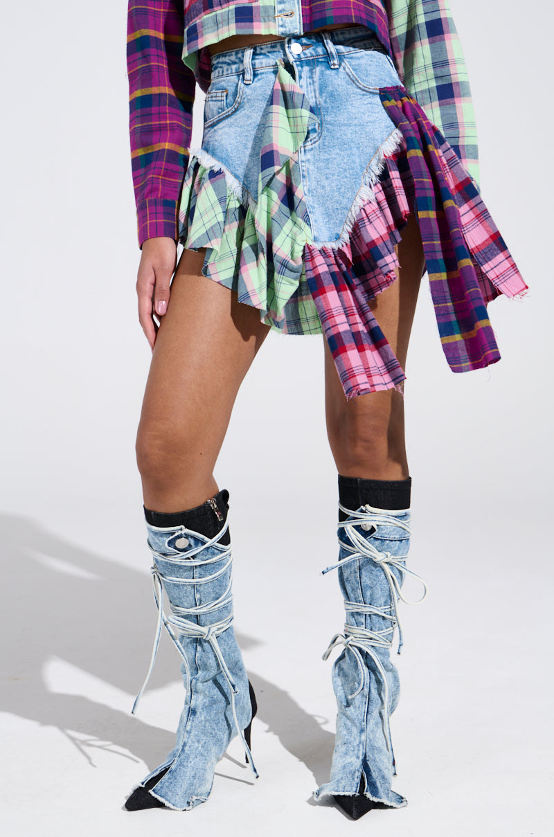 Someone sports a striking outfit featuring the THINK ABOUT ME MORE MINI SKIRT WITH PLAID DETAILS, paired with a matching plaid long-sleeve shirt. The look is completed with denim lace-up boots with pointed toes, set against a plain white backdrop.