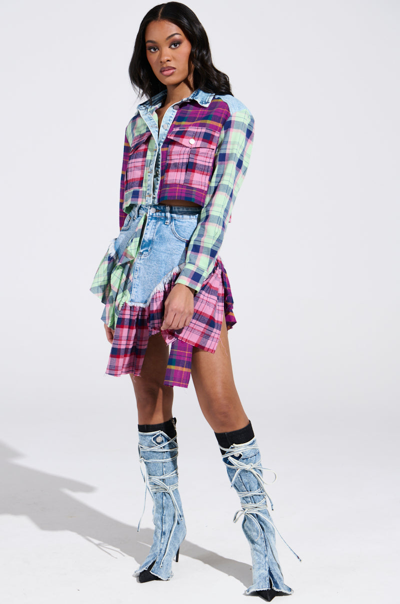 A woman exudes style and poise in her 'Think About Me More' mini skirt with plaid details, complemented by a plaid shirt dress in pink, purple, and green. She pairs these effortlessly with tall lace-up boots against a plain white background.