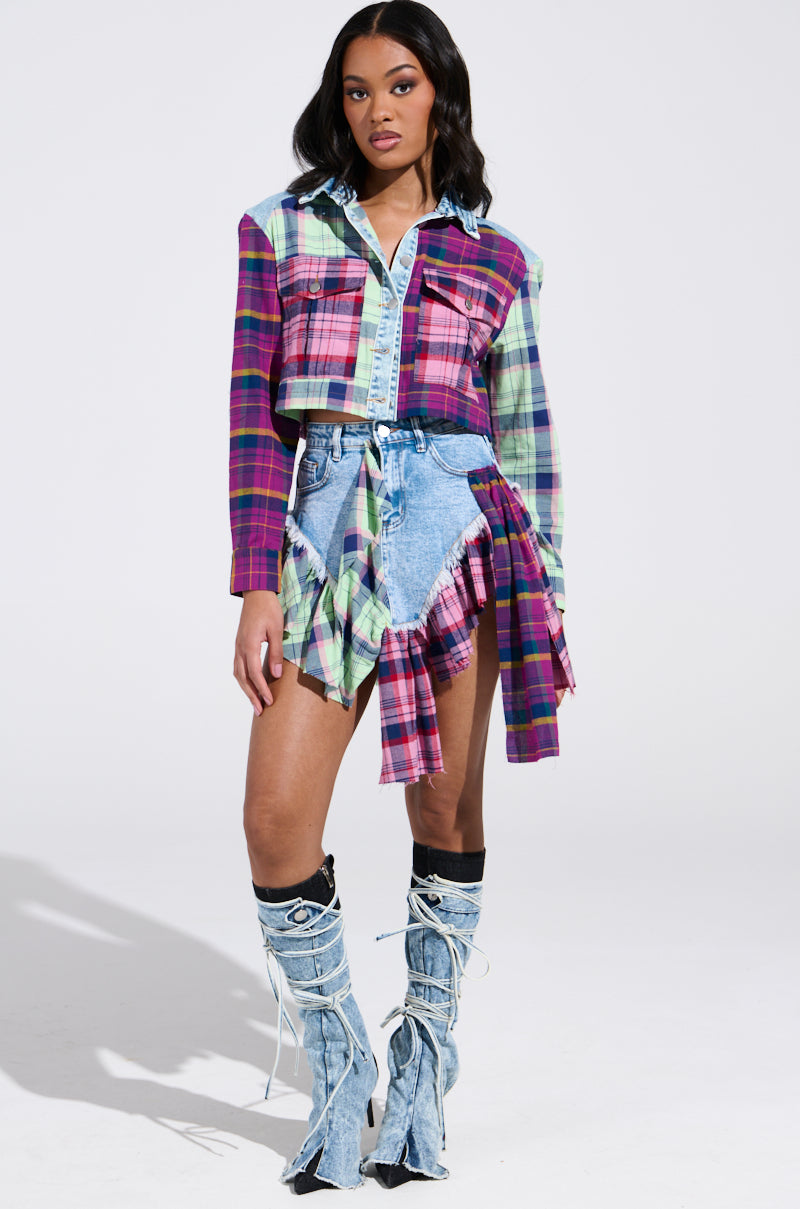 Against a plain backdrop, a person wears the THINK ABOUT ME MORE MINI SKIRT WITH PLAID DETAILS paired with asymmetrical denim shorts featuring ruffled edges. They complete the look with knee-high lace-up denim boots with frayed detailing and shoulder-length hair styled in loose waves.
