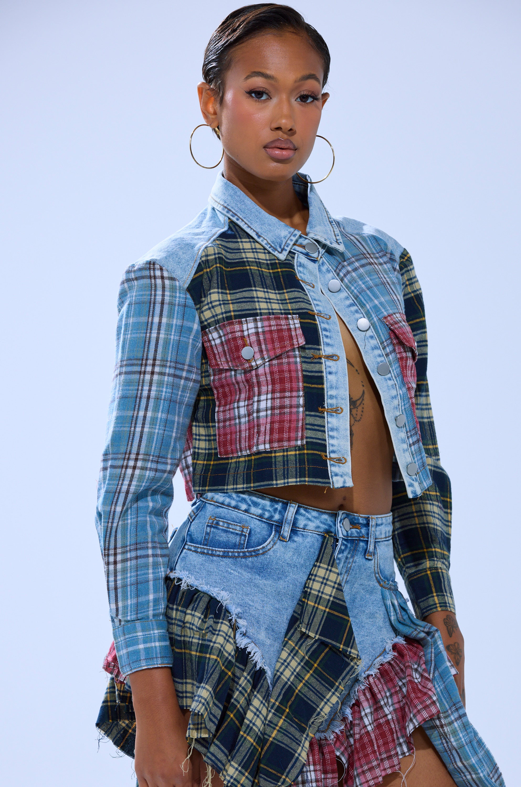 A woman in large hoop earrings wears the AUTUMN VIBES PLAID BUTTON DOWN and a matching frayed-edge patchwork skirt with a denim jacket. Her hair is slicked back as she stands confidently against a plain backdrop.
