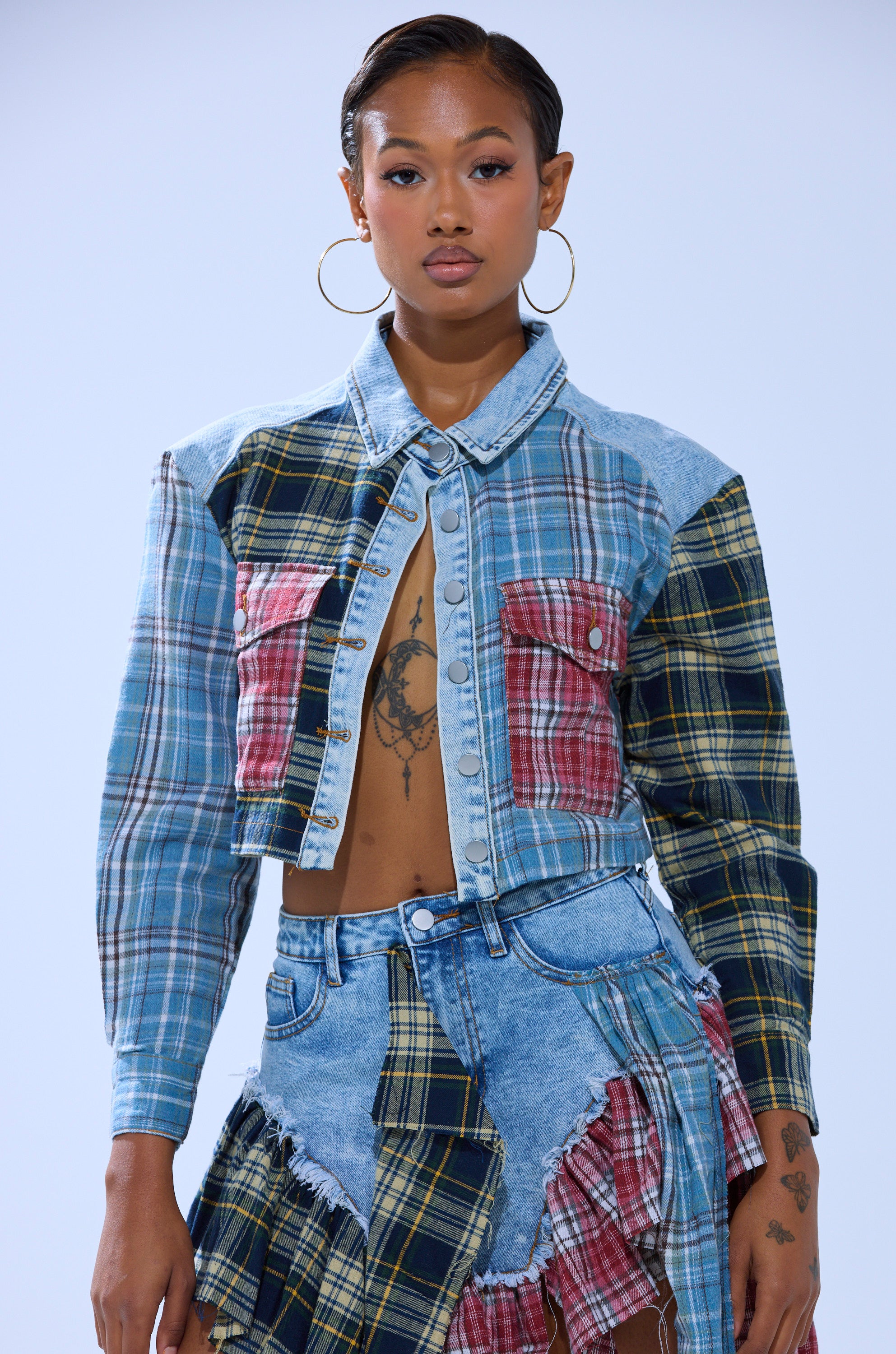 A woman with slicked-back hair and visible tattoos wears the AUTUMN VIBES PLAID BUTTON DOWN as a cropped jacket with a matching skirt, accessorized with large hoop earrings, standing against a plain background.