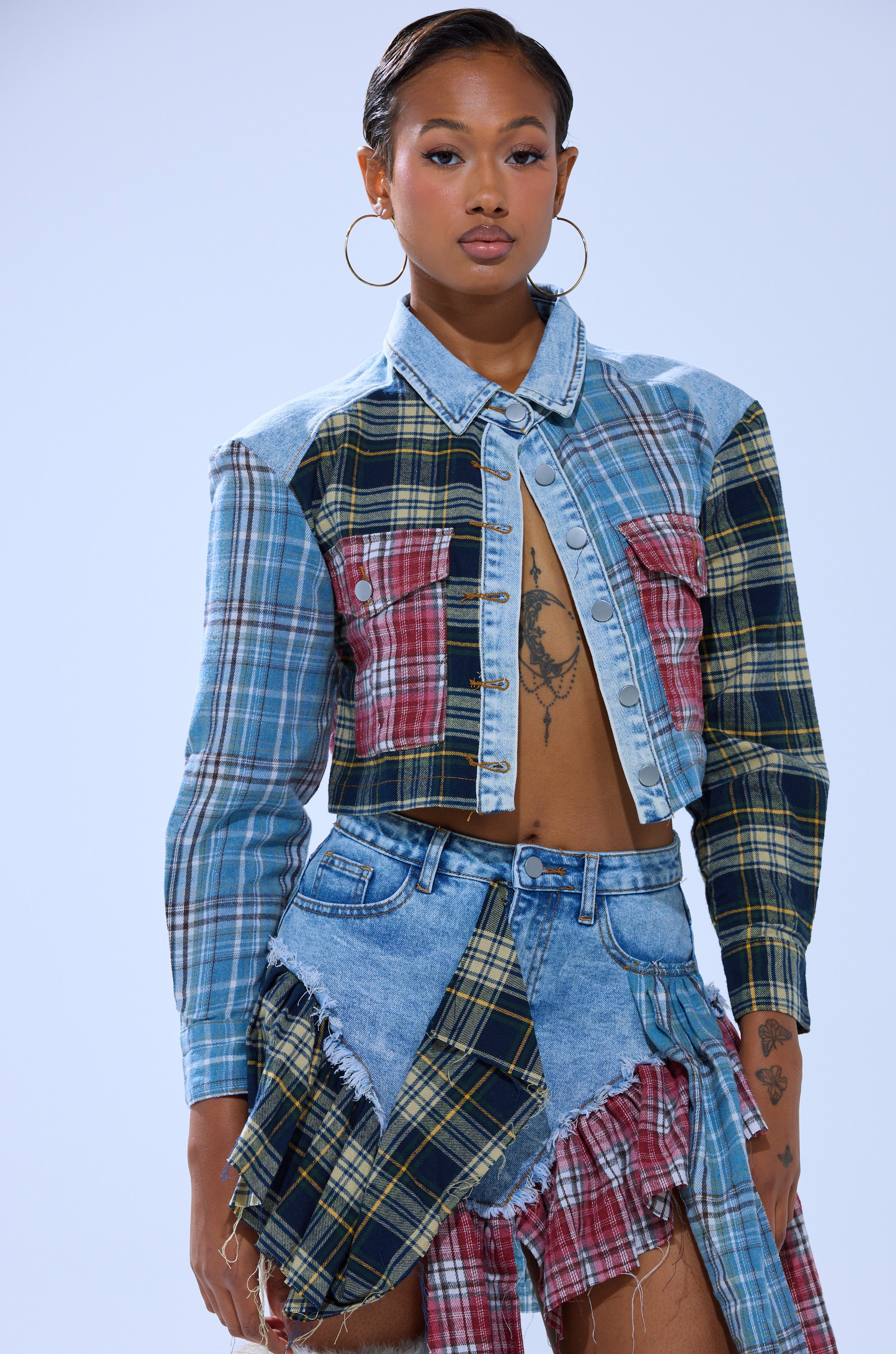 A woman wears the AUTUMN VIBES PLAID BUTTON DOWN as a crop jacket paired with a matching asymmetrical skirt, hoop earrings, and sports tattoos and a sleek hairstyle, posing against a plain light background.