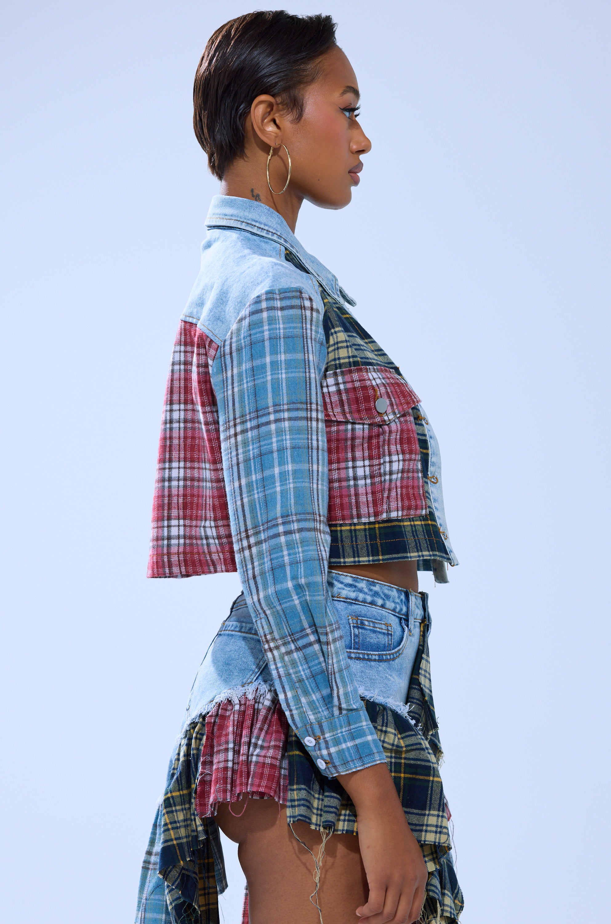 A woman with short hair and hoop earrings poses in profile against a light blue background, wearing the AUTUMN VIBES PLAID BUTTON DOWN—a cropped plaid and denim jacket paired with a matching skirt.