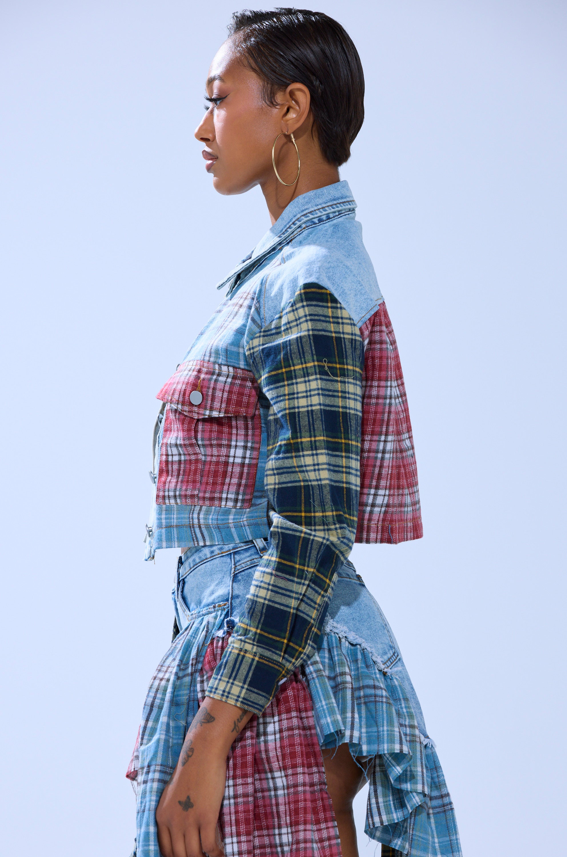 Wearing the AUTUMN VIBES PLAID BUTTON DOWN, a person with short, slicked-back hair poses in profile against a light blue background, paired with a patchwork skirt, hoop earrings, and visible hand tattoos.