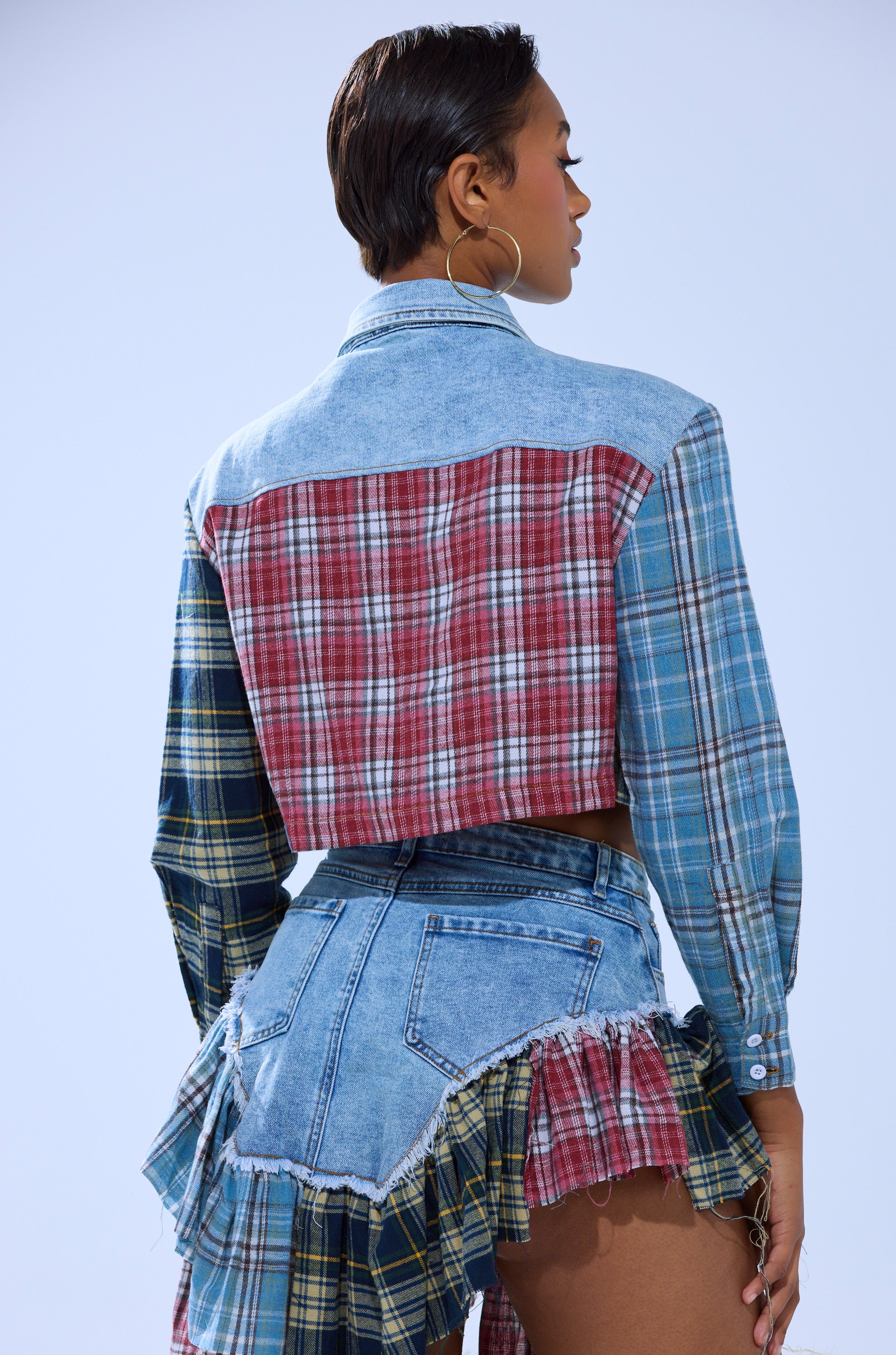 A woman with short hair faces away, wearing the AUTUMN VIBES PLAID BUTTON DOWN patchwork shirt and frayed denim shorts with plaid and denim ruffle details, set against a plain light blue background.