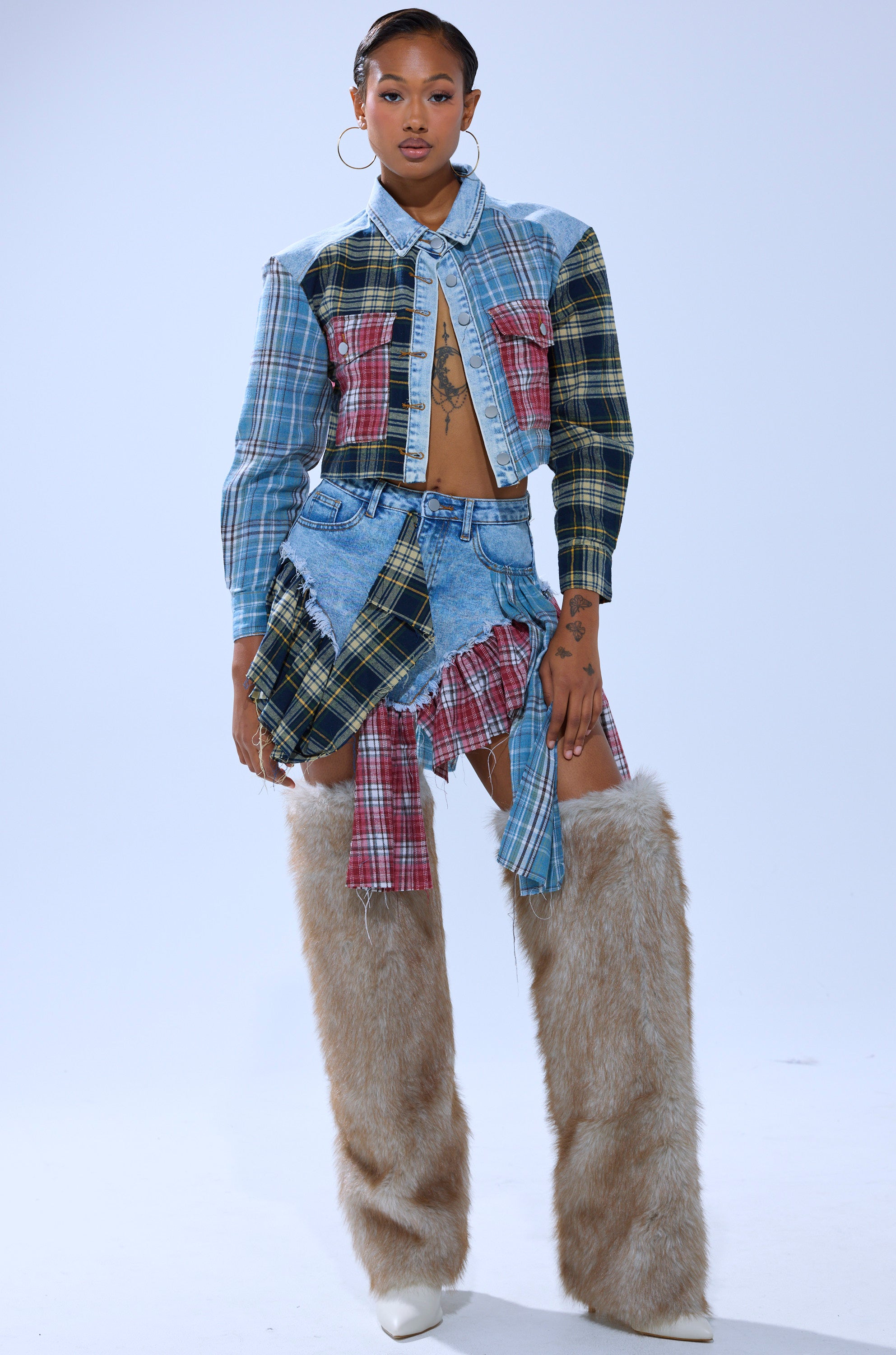 A woman showcases the AUTUMN VIBES PLAID BUTTON DOWN styled as a crop top and skirt set with asymmetrical details, tall furry beige leg warmers, white boots, hoop earrings, and visible torso and hand tattoos.