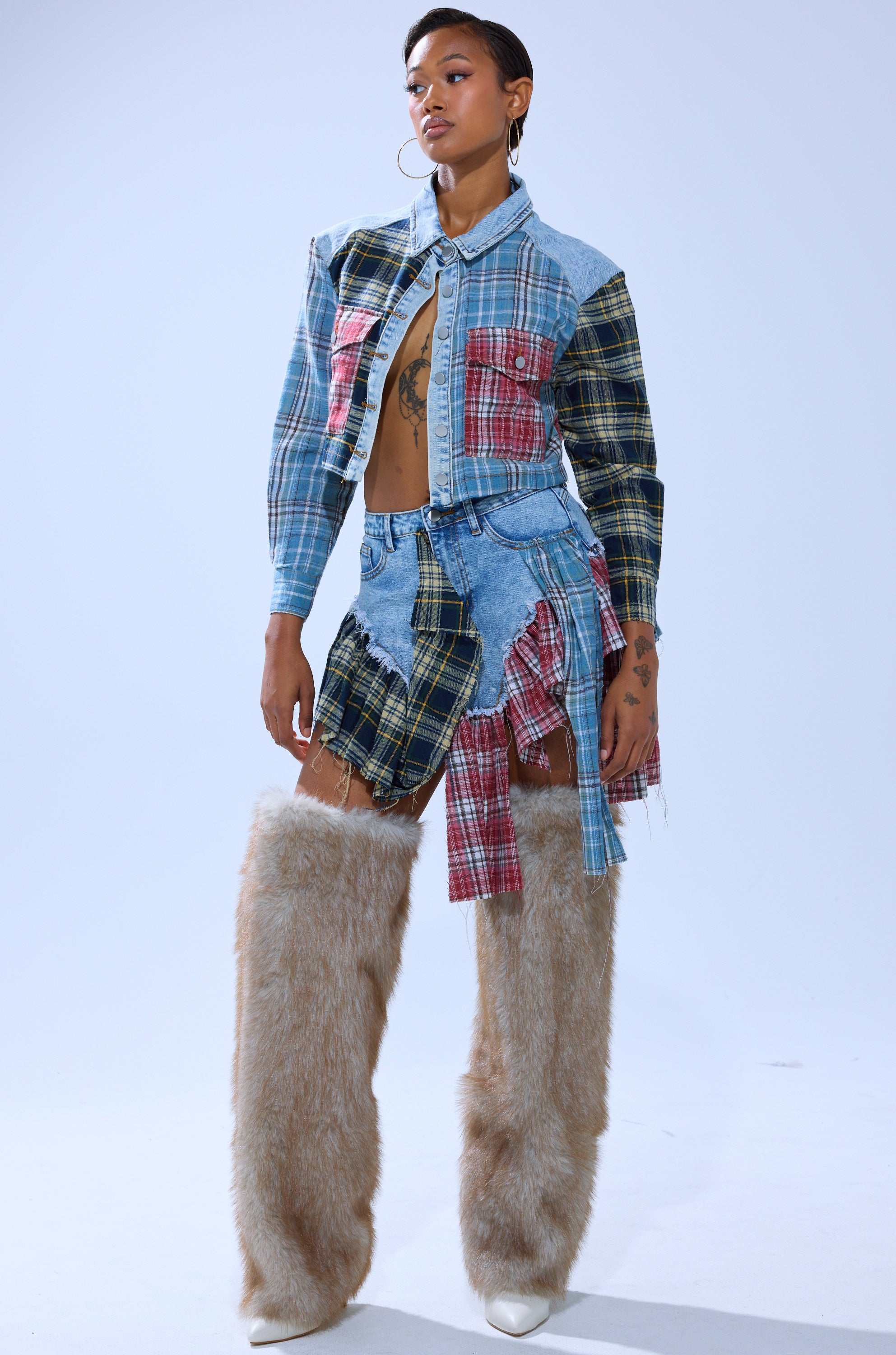 A person stands confidently in the AUTUMN VIBES PLAID BUTTON DOWN with a denim cropped jacket, skirt, large hoop earrings, and beige furry leg warmers over white shoes.