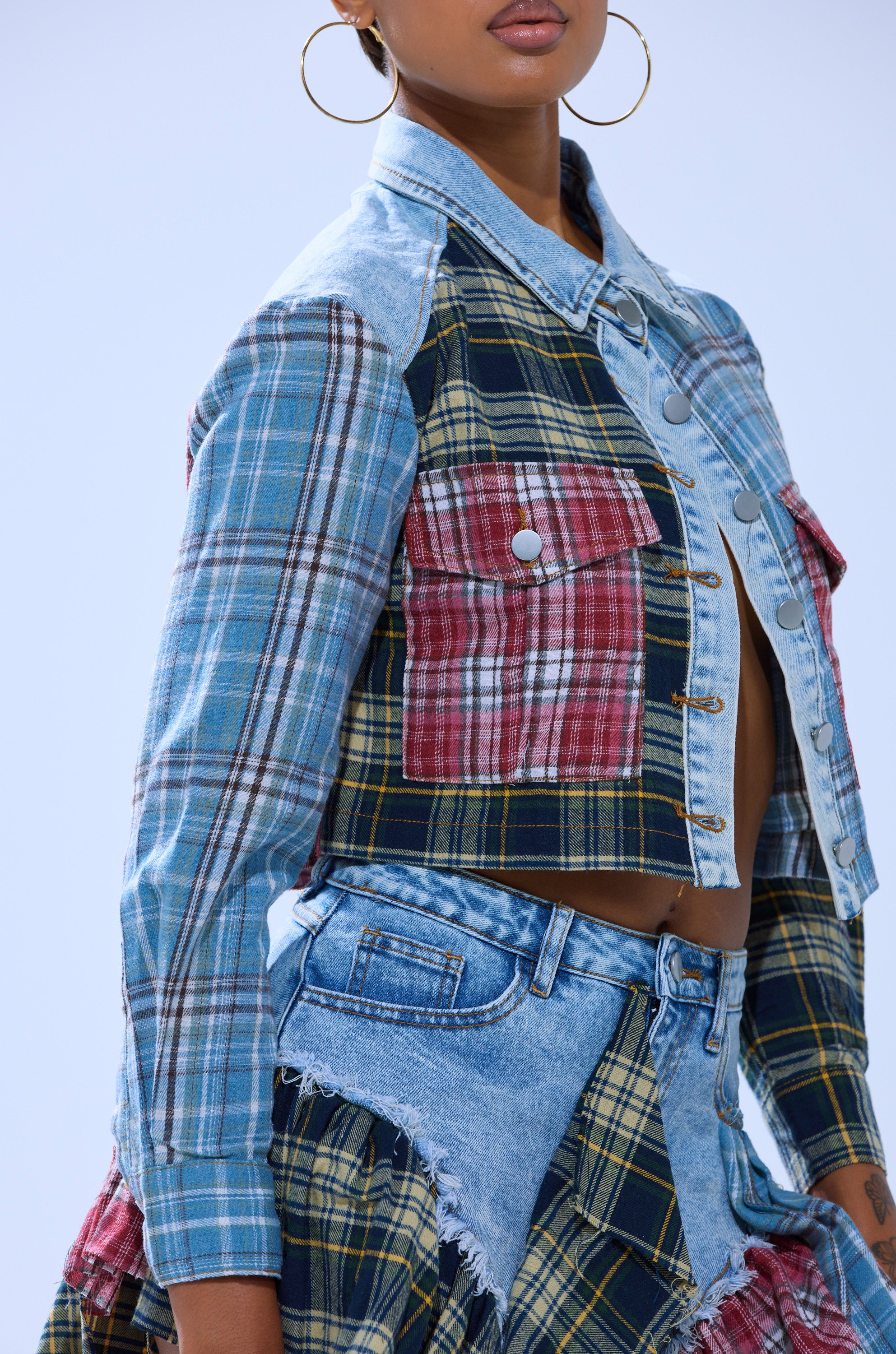 A person wears the AUTUMN VIBES PLAID BUTTON DOWN, a cropped patchwork jacket with mixed plaid and denim patterns, large red plaid pockets, paired with a matching patchwork skirt and gold hoop earrings.