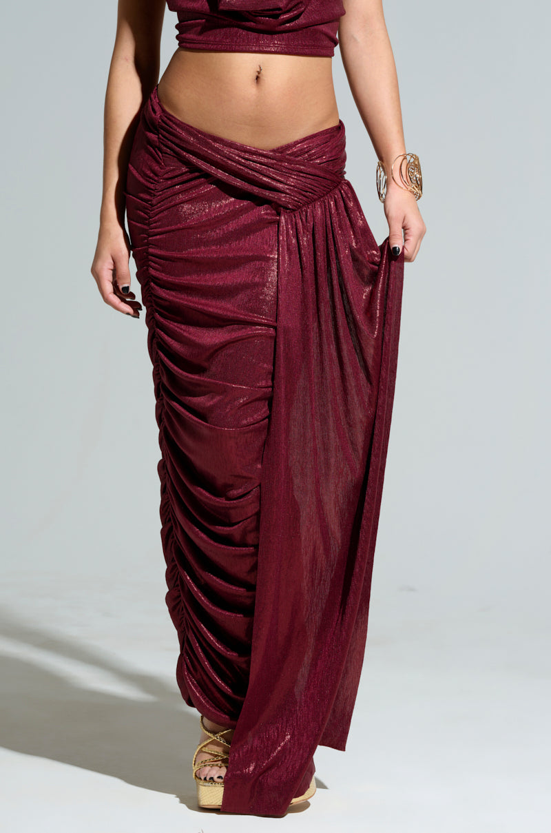 The individual is elegantly styled in the ALL OF THE GLAMOUR SLINKY MAXI SKIRT, featuring a ruched construction in burgundy, complemented by a coordinating top. They accentuate the look with a gold bracelet and metallic sandals. The glamorous skirt flows gracefully to one side against the plain gray background.