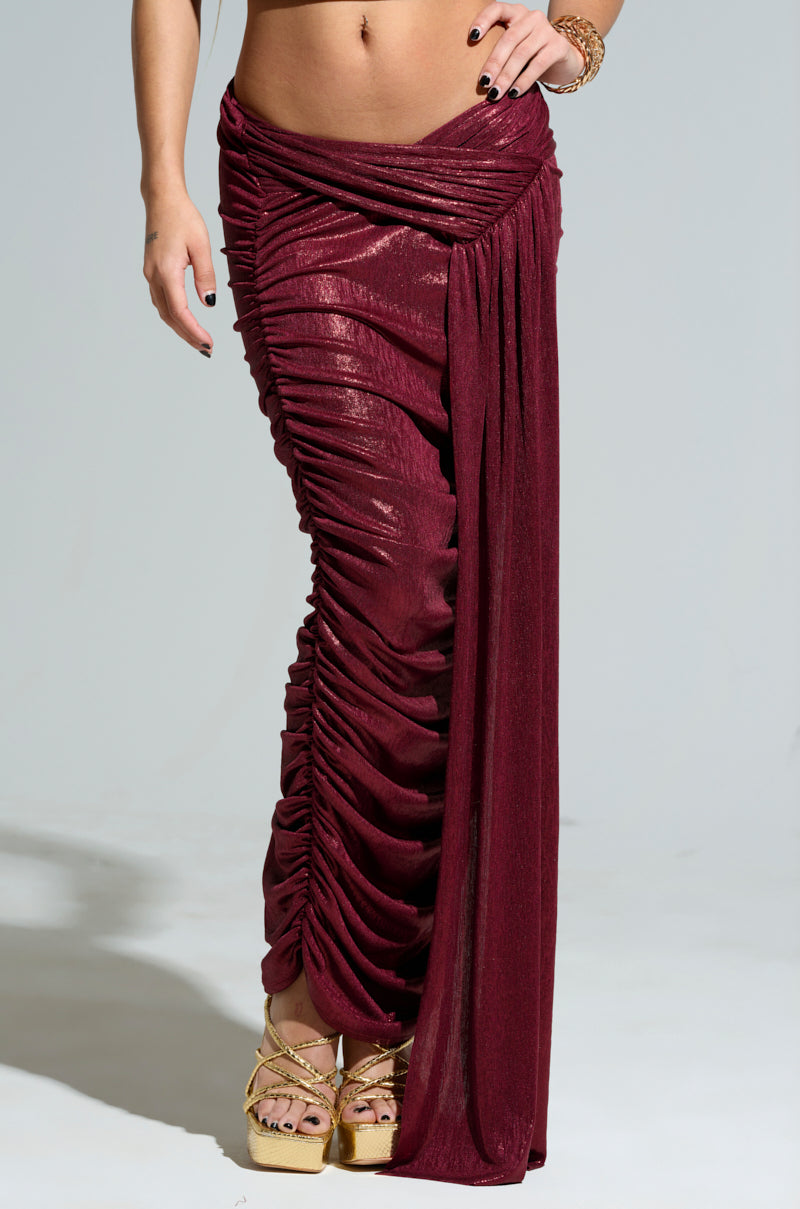 A person is wearing the ALL OF THE GLAMOUR SLINKY MAXI SKIRT in burgundy, featuring a ruched construction and a shiny texture that drapes elegantly to the floor. They are paired with gold platform sandals, and their hand is adorned with a gold bracelet. The background is a neutral gray.
