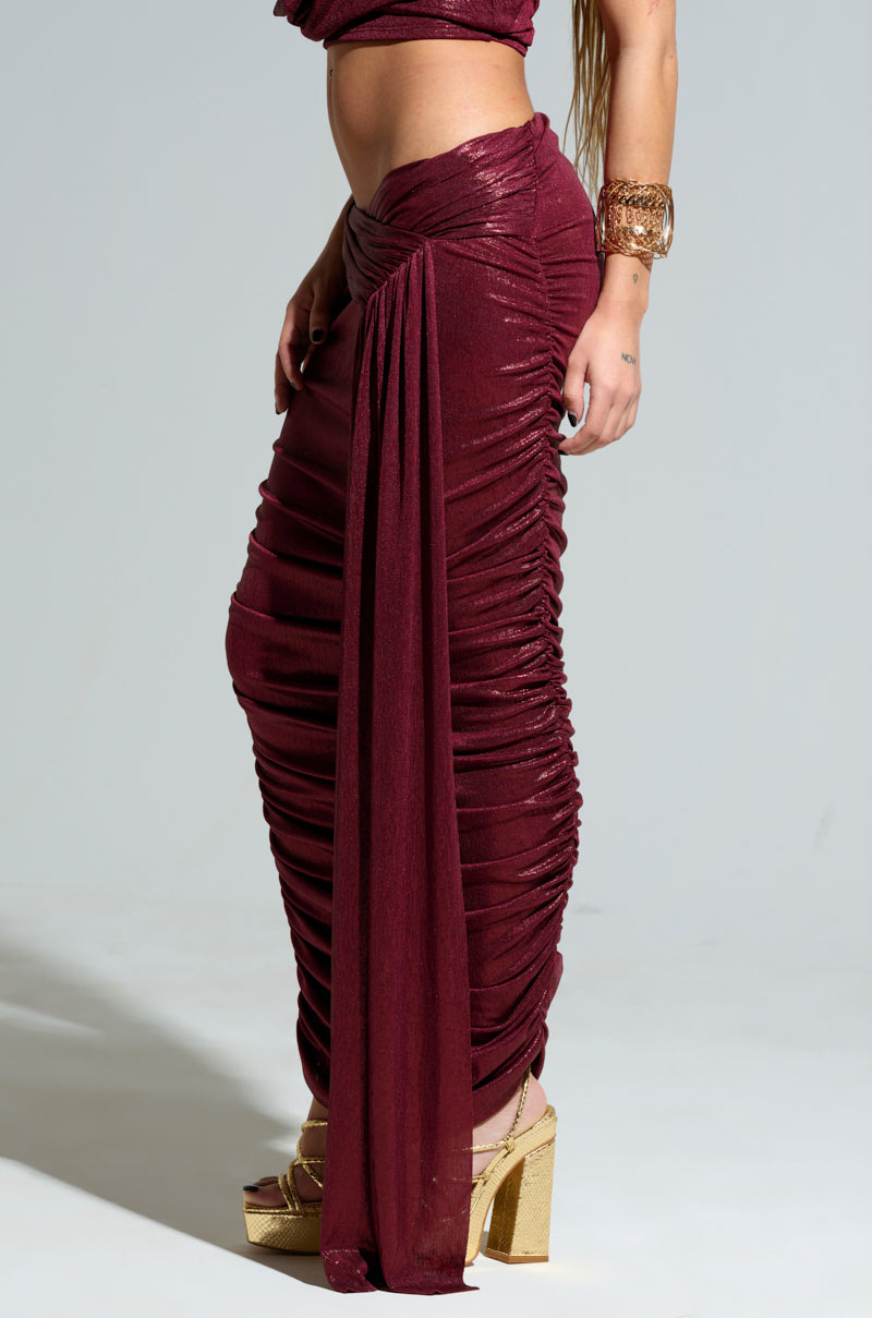 A person dressed in the "ALL OF THE GLAMOUR SLINKY MAXI SKIRT" in a shimmering burgundy hue, featuring a ruched design and side slit, stands elegantly. They complement the skirt with a matching top, a wide gold bracelet, and gold platform heels, posing gracefully against a plain background.
