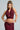 A person poses confidently, adorned in the ALL OF THE GLAMOUR SLINKY HALTER NECK TOP in a shimmering maroon color, paired perfectly with a matching skirt featuring ruched details that highlight its body-hugging silhouette. Their long braided hair is complemented by hoop earrings and several bracelets, while a tattoo adorns their arm. Their expression remains serious.