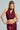 A person with braided hair wears the ALL OF THE GLAMOUR SLINKY HALTER NECK TOP, a sleeveless metallic knit in burgundy, paired with a matching skirt. They pose confidently with one hand on their hip against a plain background, showcasing their body-hugging silhouette.