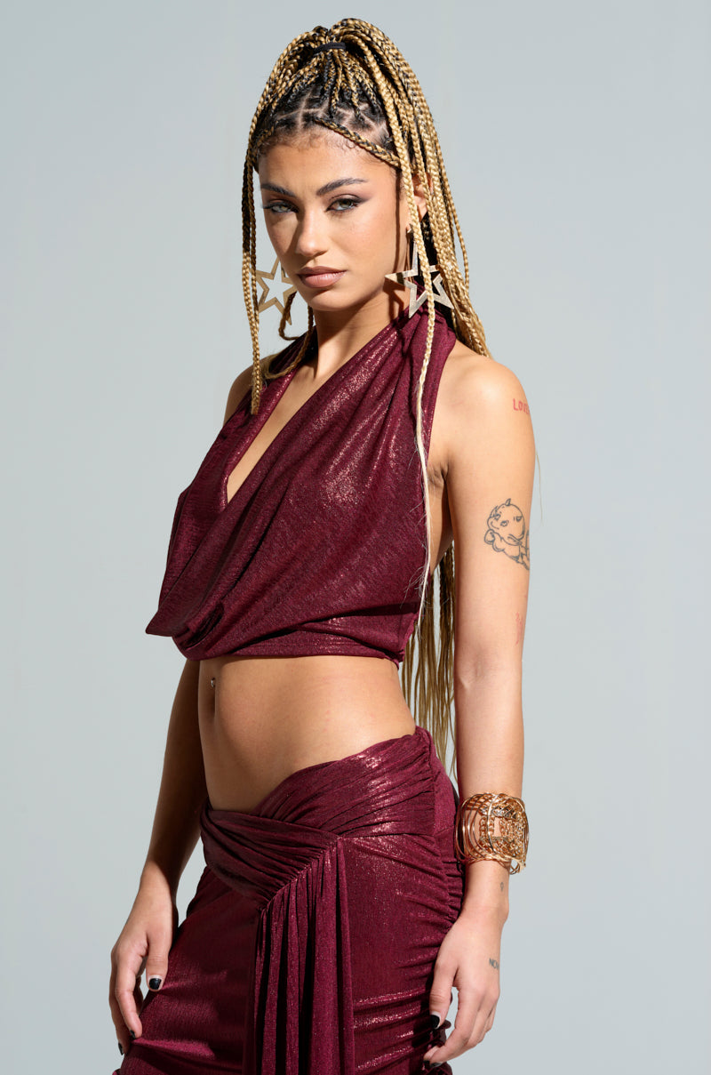 A person with long braided hair wears the ALL OF THE GLAMOUR Slinky Halter Neck Top, which is stylish and body-hugging in a sleeveless burgundy design. They accessorize with large star-shaped earrings and showcase tattoos on their arm. With a confident expression, they pose against a neutral background.