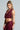A person with long braided hair wears the ALL OF THE GLAMOUR Slinky Halter Neck Top, which is stylish and body-hugging in a sleeveless burgundy design. They accessorize with large star-shaped earrings and showcase tattoos on their arm. With a confident expression, they pose against a neutral background.