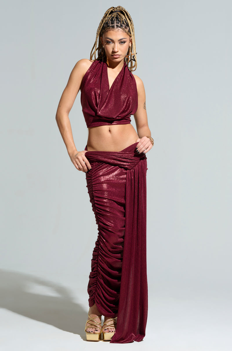 Wearing the ALL OF THE GLAMOUR SLINKY HALTER NECK TOP in maroon with a shimmering metallic knit, paired with a matching ruched skirt featuring a draped accent, an individual stands confidently against a plain background. Their braided hair complements the ensemble as they pose with one hand elegantly resting on their hip.