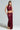 Wearing the ALL OF THE GLAMOUR SLINKY HALTER NECK TOP in maroon with a shimmering metallic knit, paired with a matching ruched skirt featuring a draped accent, an individual stands confidently against a plain background. Their braided hair complements the ensemble as they pose with one hand elegantly resting on their hip.