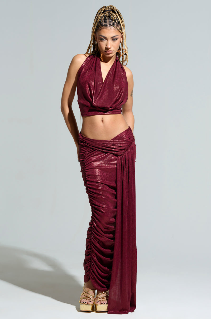 A woman with braided hair poses confidently in a shimmering burgundy ensemble, showcasing the ALL OF THE GLAMOUR SLINKY HALTER NECK TOP paired with a ruched skirt that accentuates her curves. She stands against a plain background, completing her look with high platform sandals.