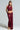 A woman with braided hair poses confidently in a shimmering burgundy ensemble, showcasing the ALL OF THE GLAMOUR SLINKY HALTER NECK TOP paired with a ruched skirt that accentuates her curves. She stands against a plain background, completing her look with high platform sandals.