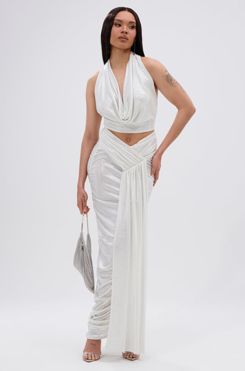 A woman poses in the ALL OF THE GLAMOUR SLINKY MAXI SKIRT IN WHITE, featuring ruched detail and a sash, paired with a shimmery white halter top and silver handbag. She wears strappy heels and stands against a plain white background. 2