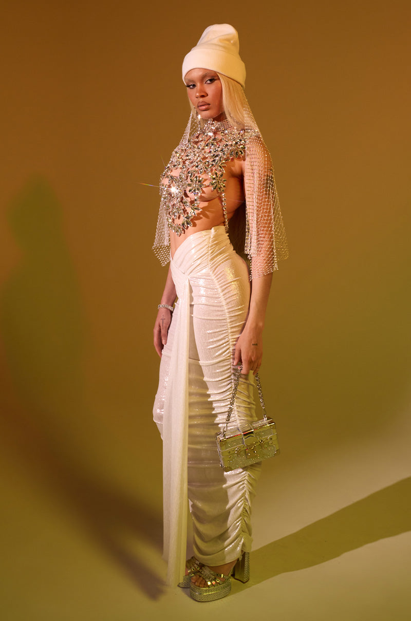 Against a warm-toned backdrop, a person wears the ALL OF THE GLAMOUR SLINKY MAXI SKIRT IN WHITE with a bejeweled top, white head wrap, mesh veil, platform heels, statement earrings, and holds a silver handbag. 1