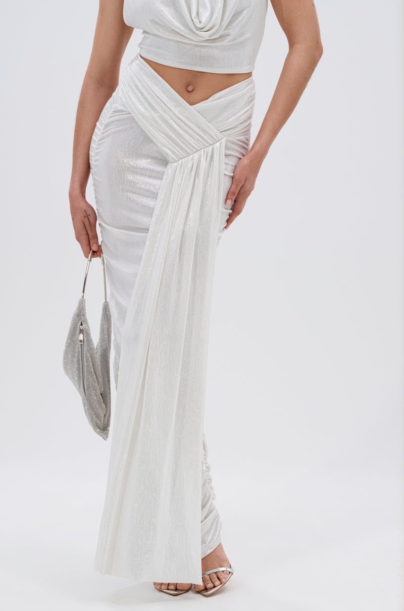 A woman models the ALL OF THE GLAMOUR SLINKY MAXI SKIRT IN WHITE, paired with a matching sleeveless top and silver clutch. She stands in open-toe heels before a plain light background, her face out of frame. 5