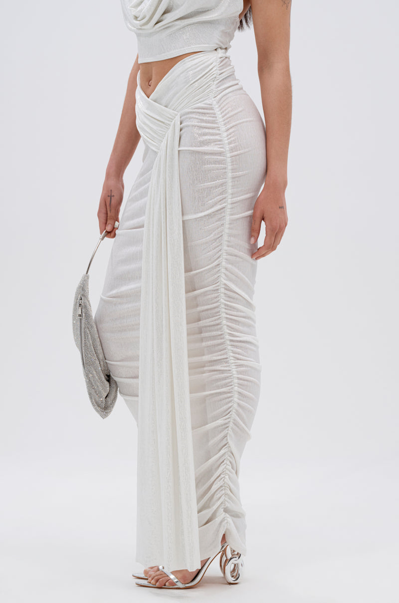 A woman wears the ALL OF THE GLAMOUR SLINKY MAXI SKIRT IN WHITE—a sheer, ruched skirt with an asymmetrical V-line waist and draped sash—paired with a matching sleeveless top, silver clutch, and strappy heeled sandals against a white background. 7
