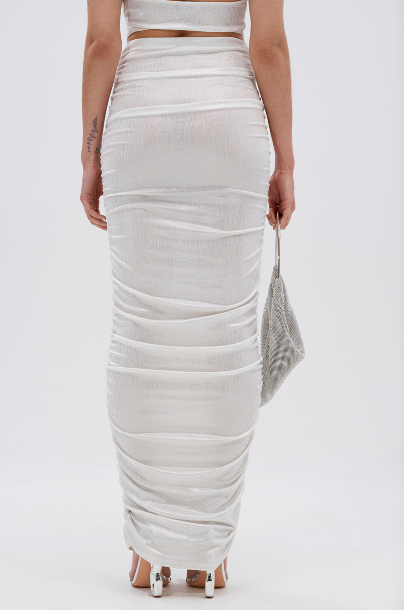 Wearing the ALL OF THE GLAMOUR SLINKY MAXI SKIRT IN WHITE, a person stands with white heeled sandals and a sparkly silver handbag, their arm tattoo visible against a plain light background. Viewed from the back, the skirt’s ruched silhouette is featured. 8