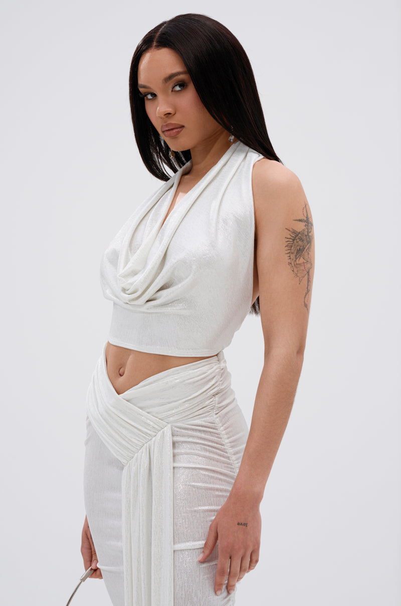 A woman with long straight dark hair wears the ALL OF THE GLAMOUR SLINKY HALTER NECK TOP IN WHITE, a sleeveless crop top with a plunging cowl neckline, paired with a matching high-waisted wrap skirt. She has a tattoo on her upper left arm and stands against a light background.