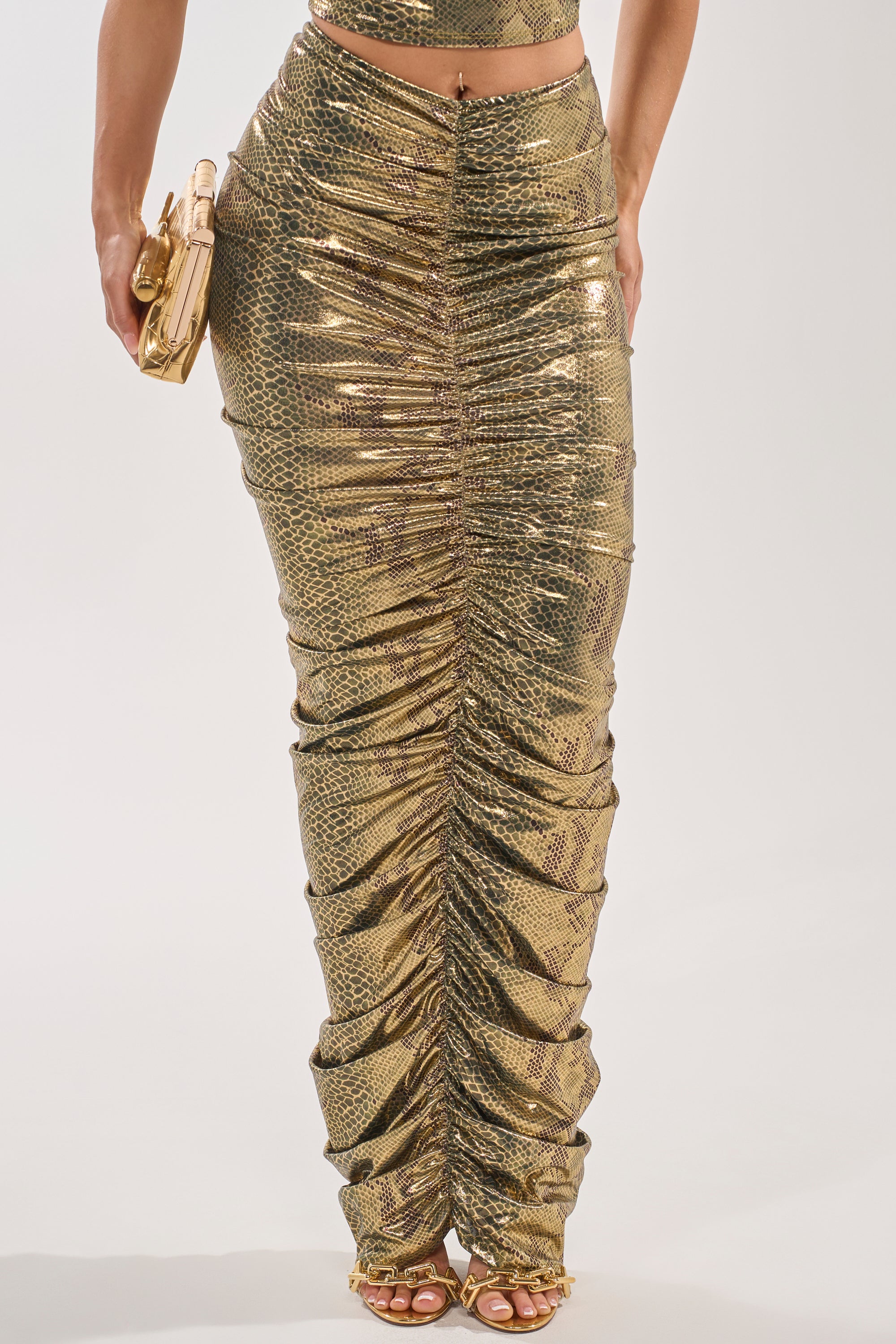 Wearing the SNEAKY LITTLE SNAKE MAXI SKIRT, a high-rise metallic snakeskin skirt, paired with a matching crop top, gold clutch, and strappy gold heels. 2