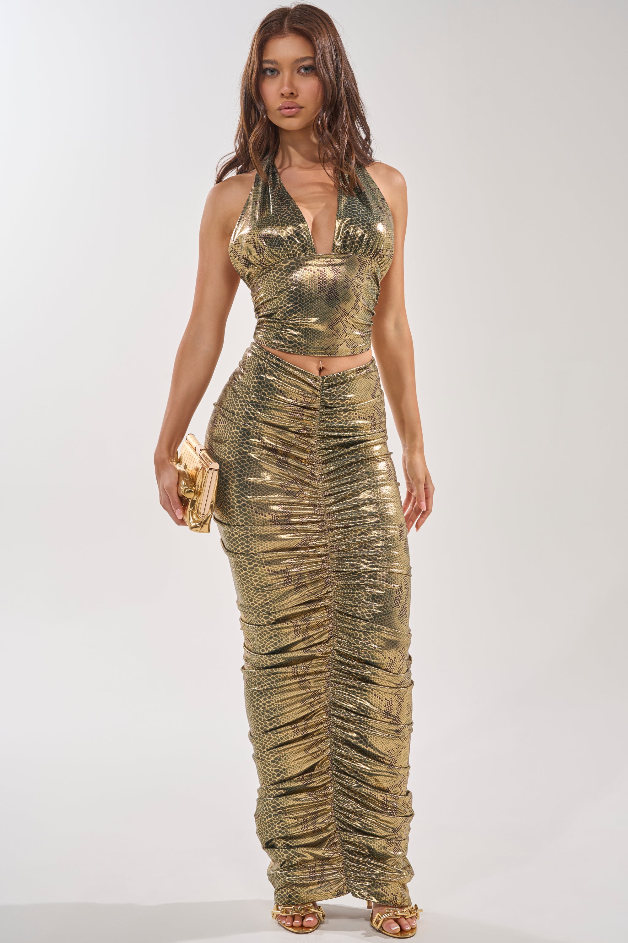A woman poses against a plain background in the SNEAKY LITTLE SNAKE HALTER TOP with a cropped fit, paired with a matching ruched skirt, gold clutch, and gold heels for an effortlessly sultry look. 5