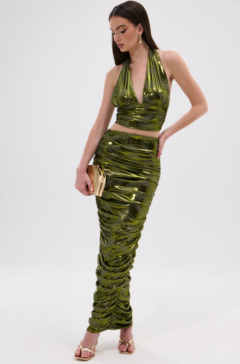 A woman stands against a white background wearing the TAYLOR METALLIC RUCHED MAXI SKIRT with a metallic green halter crop top, gold heels, gold clutch, earrings, and long brown hair, posing stylishly. 0