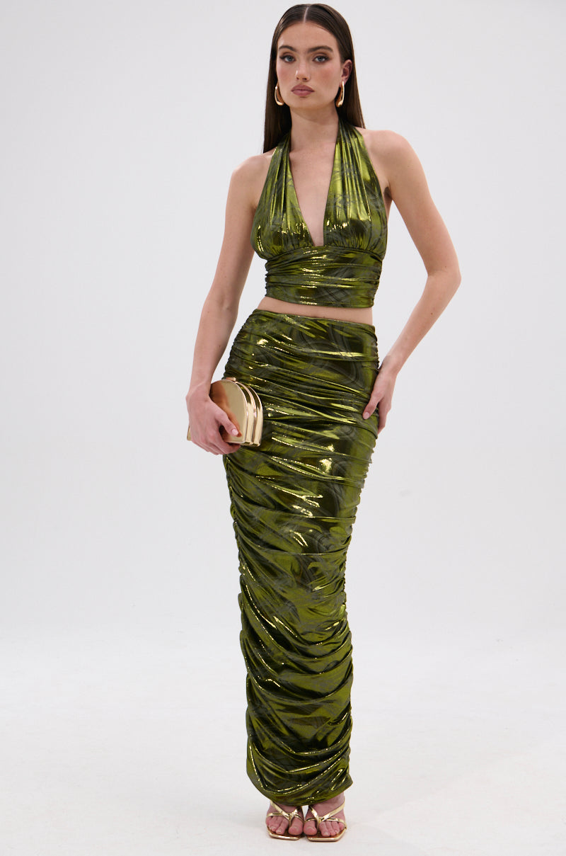A woman wearing the TAYLOR METALLIC HALTER TOP IN GREEN with a matching maxi skirt, gold clutch, hoop earrings, and long straight hair stands against a plain white background. 4