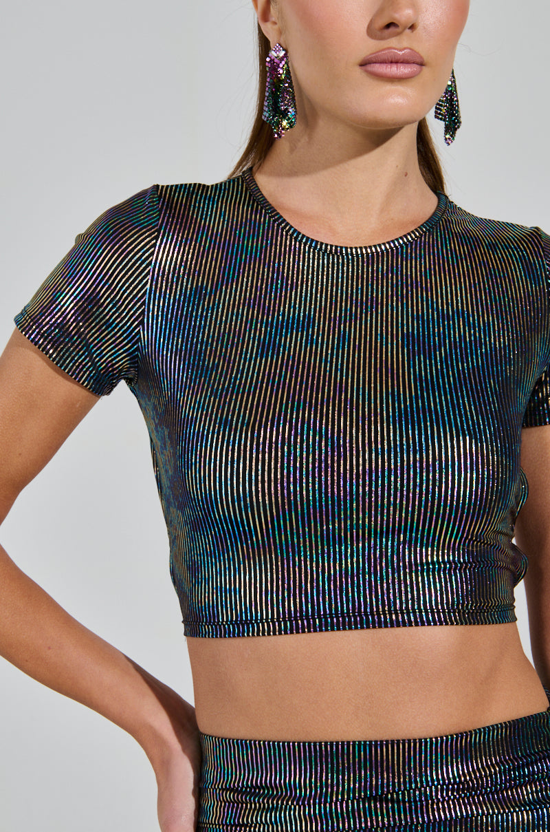 A person wearing the ZEON TANK WITH TIE IN BACK, featuring a shimmering iridescent striped design and an open back, paired with matching earrings. They have their hand on their hip, standing against a plain background.