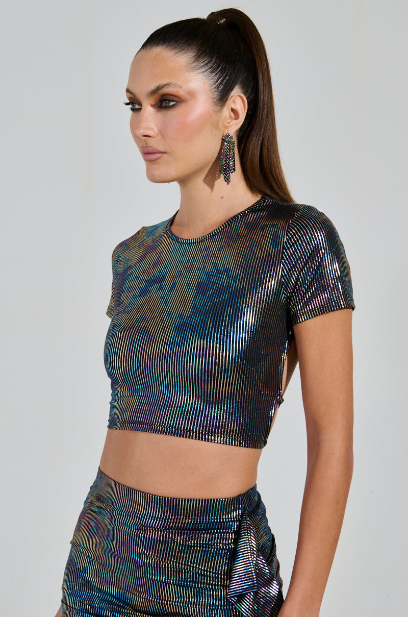 A woman with a ponytail wears the ZEON TANK WITH TIE IN BACK and matching skirt, featuring shimmering iridescent stripes. Her large dangling earrings accentuate the open-back design as she gazes to the side, showcasing a stunning holographic effect with its light-reflecting colors.