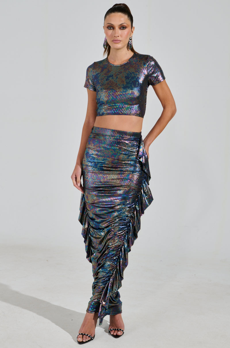 A person showcases the ZEON TANK WITH TIE IN BACK, an iridescent crop top with open back design, paired with a ruffled floor-length skirt. They complement the look with black high-heeled sandals and sleek, pulled-back hair against a plain white background.