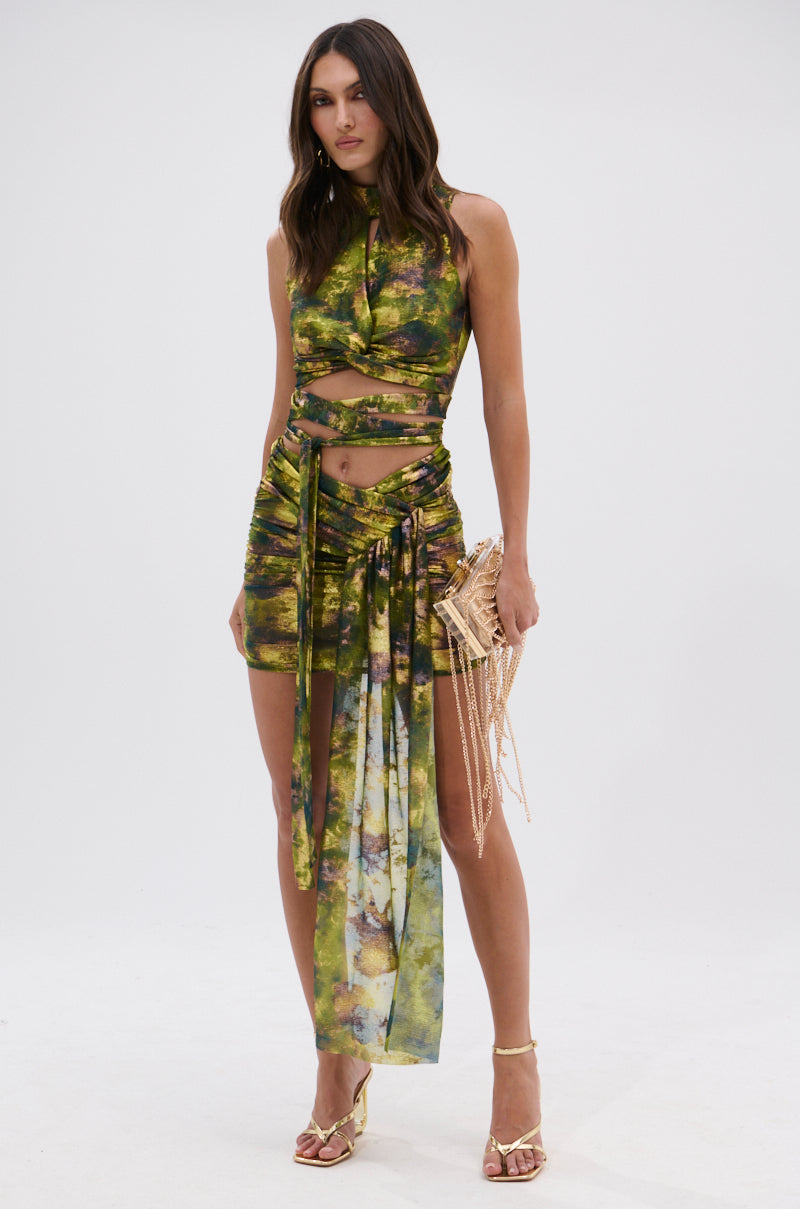 A woman wears the SEXY CAN I MINI SKIRT, an iridescent green piece with a high slit and draped fabric, paired with a green and yellow abstract crop top, gold beaded bag, and gold heels against a plain white background. 3