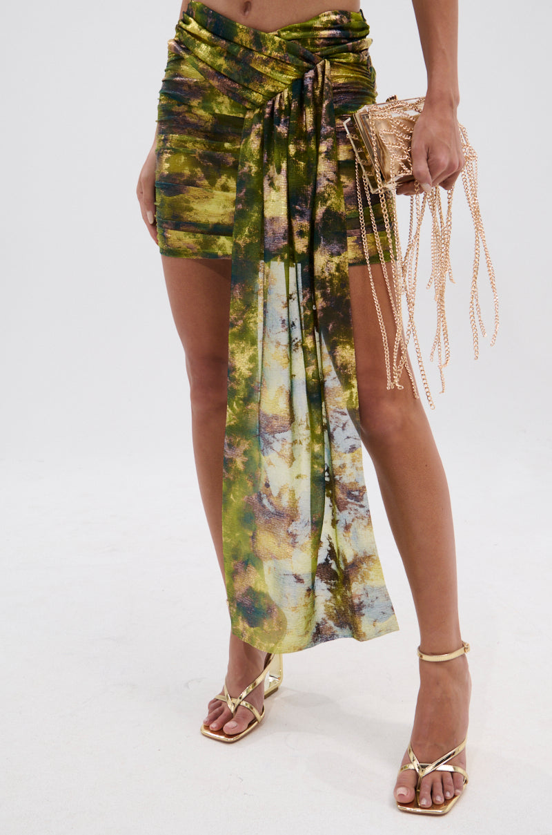 A person wearing the SEXY CAN I MINI SKIRT in green and yellow, gold strappy heels, and holding a small gold handbag with chain fringe, stands against a plain white background. 4