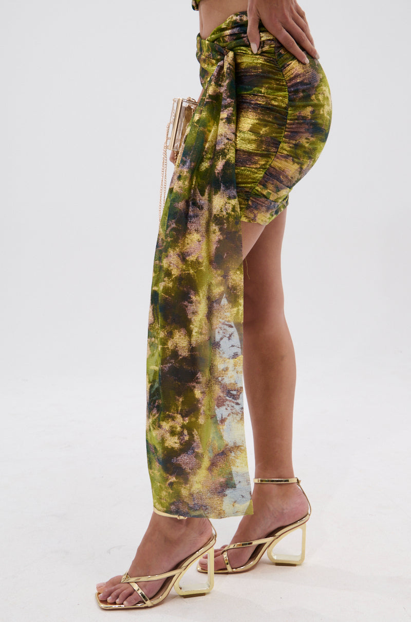 A person wearing the SEXY CAN I MINI SKIRT, featuring green and brown iridescent fabric with a long side tie, paired with gold strappy high-heeled sandals, stands against a plain white background. 5
